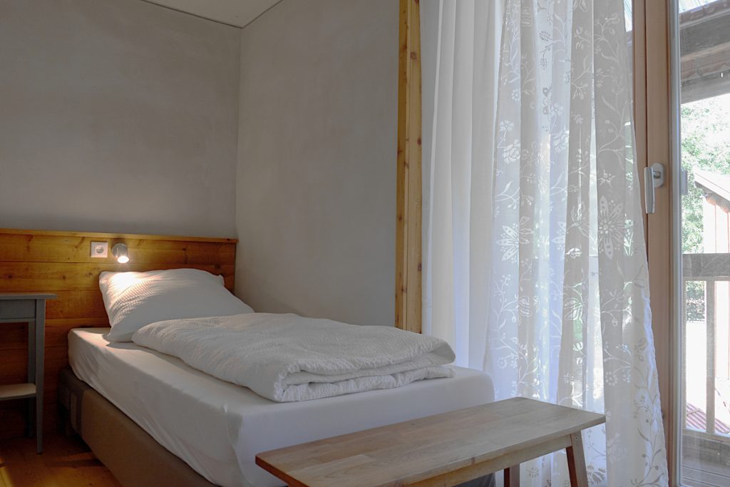 Schlafzimmer mit weißer Bettwäsche, Holzbank und Ausgang zum Laubengang.