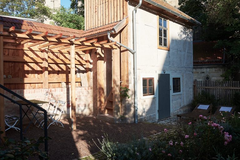 Das Ferienhaus, kleines zweistöckiges Gartenhaus mit hellgrauer Fachwerkfassade, dunkelgrauer Tür und braunen Fenstern.