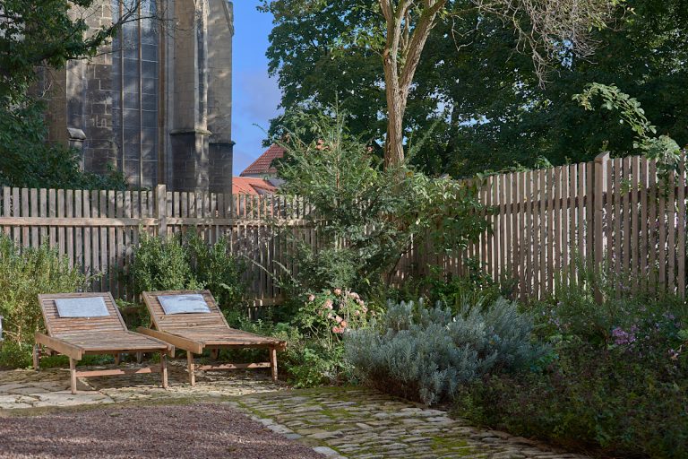 Das Ferienhaus, kleiner Garten bepflanzt mit Gehölzen und Stauden, daneben zwei hölzerne Sonnenliegen.