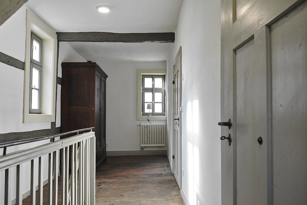 Flur mit weißen Wänden, die Türen, Treppen, Fenster und Fachwerk in unterschiedlichen Grautönen, der Holzfußboden braun.